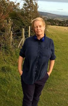 the full colour pic shows the author standing outside, in a dark blue shirt rolled up to the elbows, both hands in the pockets of her dark trousers. Her hair is blonde and blown to one side. Her face is calm and serene, but not smiling. She's looking straight at the viewer. Behind her are the rolling fields and hills of the South Downs, as well as a fence with some trees behind it. 