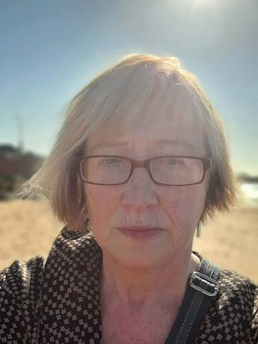 Full head and shoulders photo of author, with short blonde hair flying in a breeze and a golden field behind her. She's wearing red glasses and looks pink with the sun.