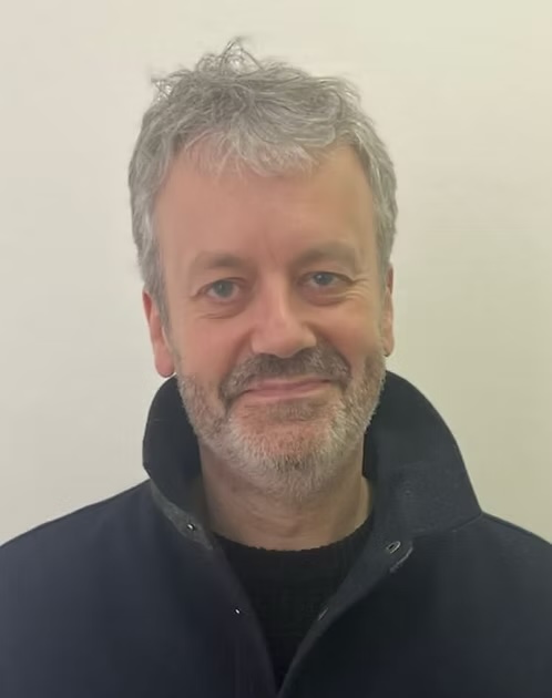 Head and shoulders photo of a male poet with short grey hair and grizzly beard growth. He is grinning.
