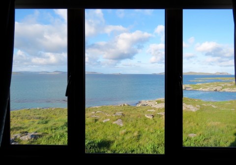 Photograph of the Hebridean coast taken through the window of Ron King's holiday house. It is divided into three sections with black frames, and is a horizontal rectangle. Through the window is amazingly turquoise see and fantastically green coastal stretches. In the sky fluffy white clouds. It looks fabulous.