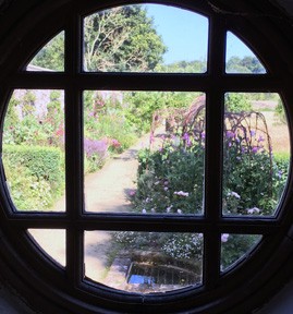 Photo shows a circular window with a lattice pattern, though which a rather lovely garden can be glimpsed with trellises and pink trailing flowers.