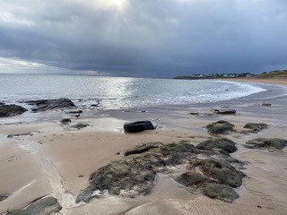 The photograph shows an empty beach, a calm sea with a lacy frill of foam touching the sand. The sky is deep grey cloud, but the sun is coming through a white gap in the middle, so that the sea has a pathway of bright light. There are low roocks in the sand. No people. It looks cold.