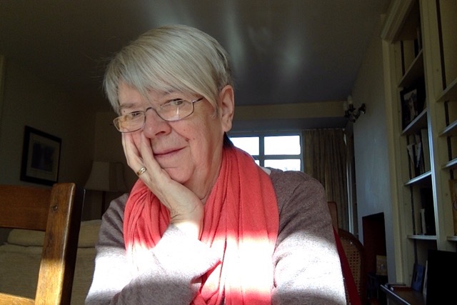 This is the poet with chin in her right hand and head slightly to one side. She is wearing glasses and has short white-grey hair. Behind her we can see most of a window, and in front of her something (perhaps another window) is throwing a wide band of light across her forearms and chest. She is wearing a salmon pink scarf looped around her next. To her left are ceiling high bookshelves. 