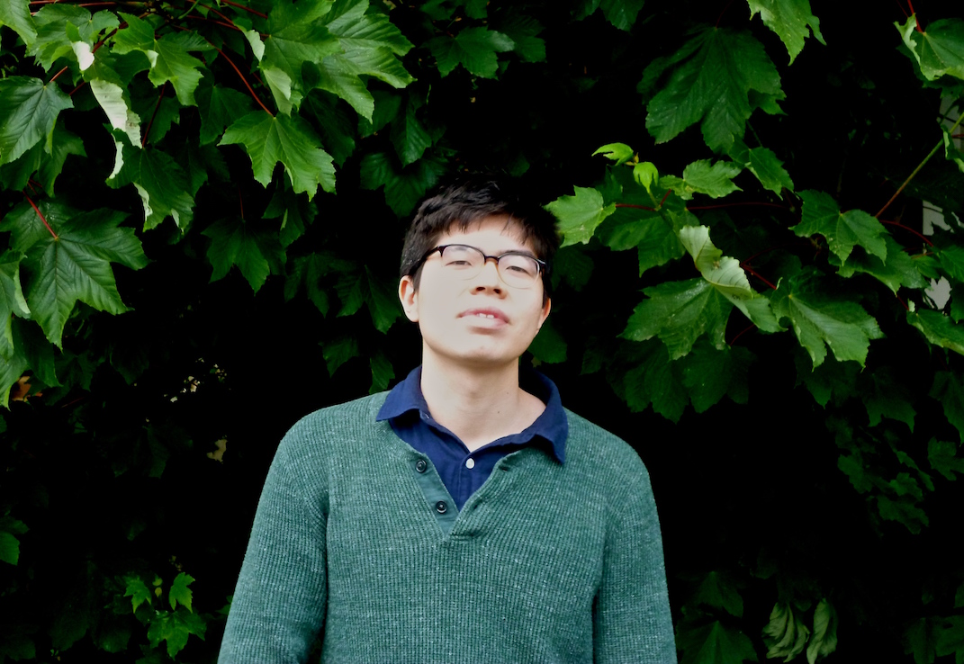 Head, torso and shoulders of Will standing against thick green foliage. He is wearing a green jumper, with an open necked dark blue shirt. Hair is dark, wears glasses, head angled up the way appraisingly, a relaxed but serious expression. He is in his early twenties to look at him, and the Indonesian heritage is obvious in his face.