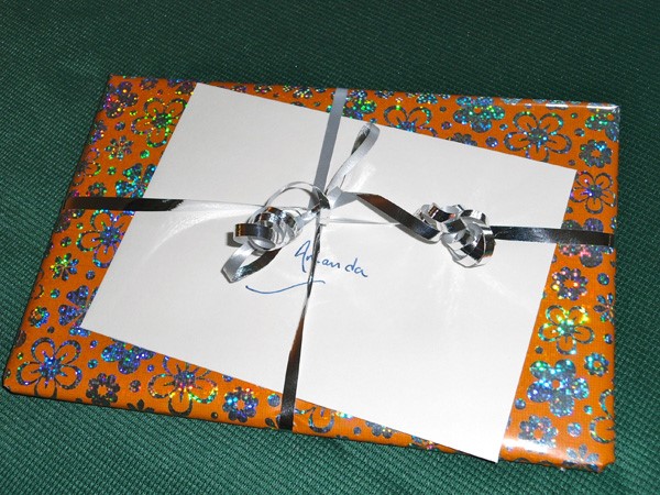 A gift wrapped in sparkly orange and silver paper with a flower motif. A whilte envelope is tucked under its silver ribbon.